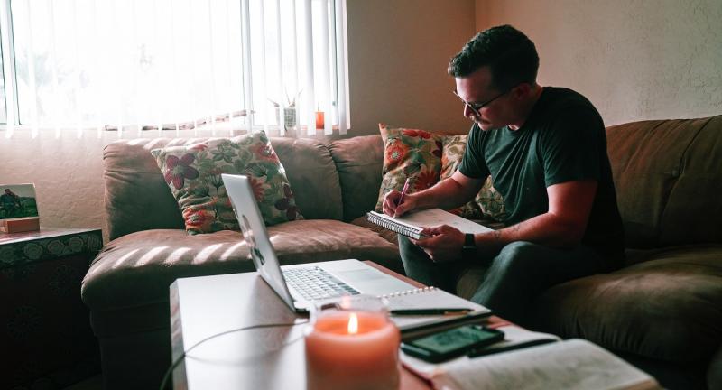 Man writing in notebook with laptop open while sitting on couch in lounge room