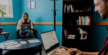 Two people smiling while working on laptops across the room from one another