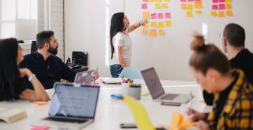 Woman pointing to post it notes on whiteboard in front of group of people at work