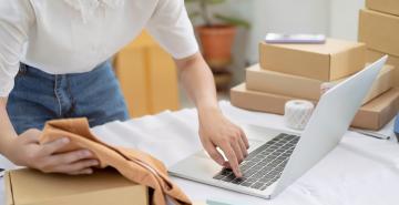 Woman on laptop near boxes on table while packaging up an online order